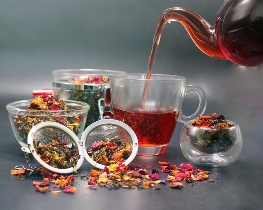 تصویری از یک قوری شیشه ای که دارد درون لیوان دمنوش سرخ رنگ شفاف میریزد و اطرافش چندین گیاه و چای است